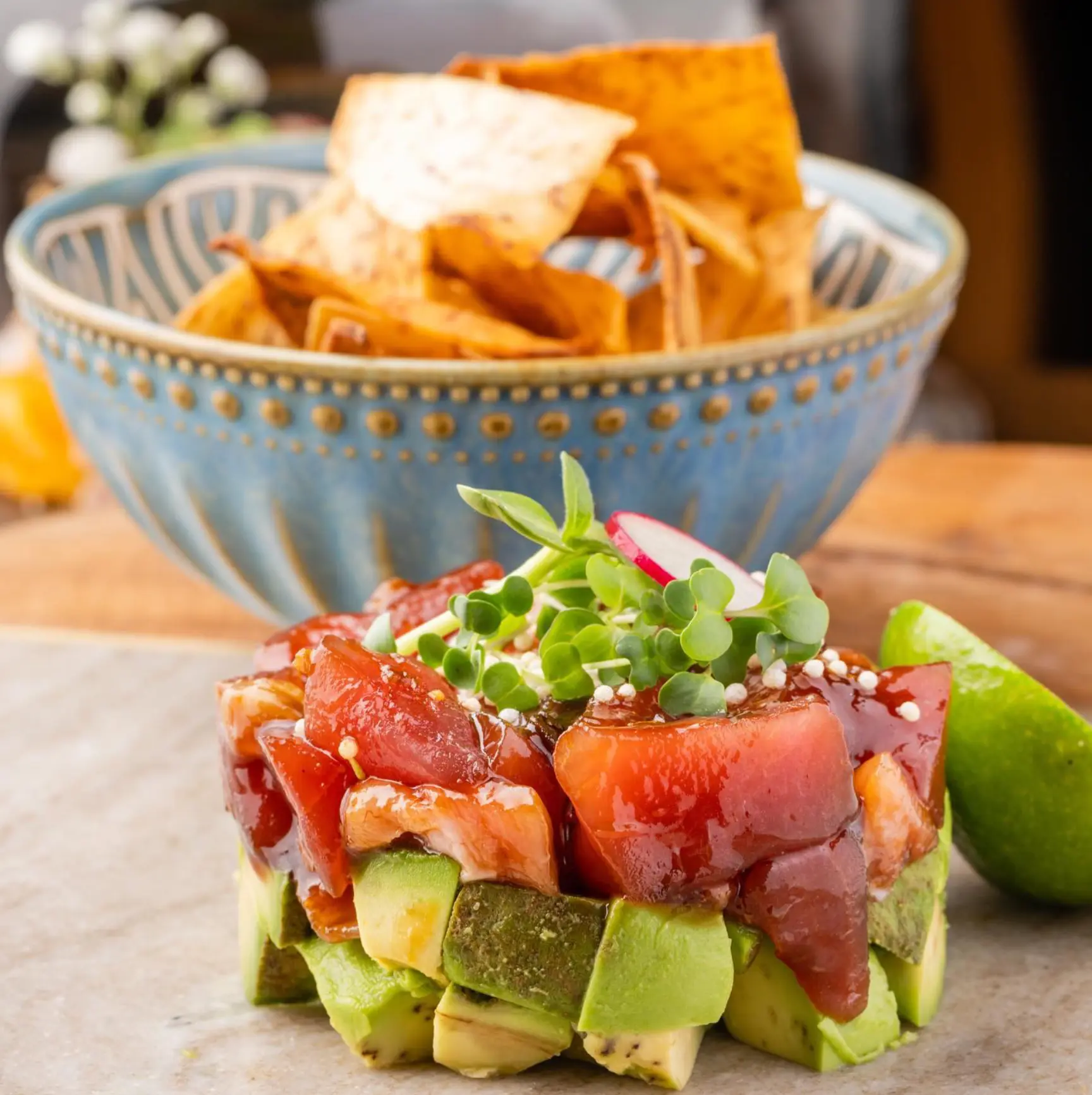 Tuna tartare tower over avocado with microgreens and a lime wedge, beside a blue bowl of crispy chips