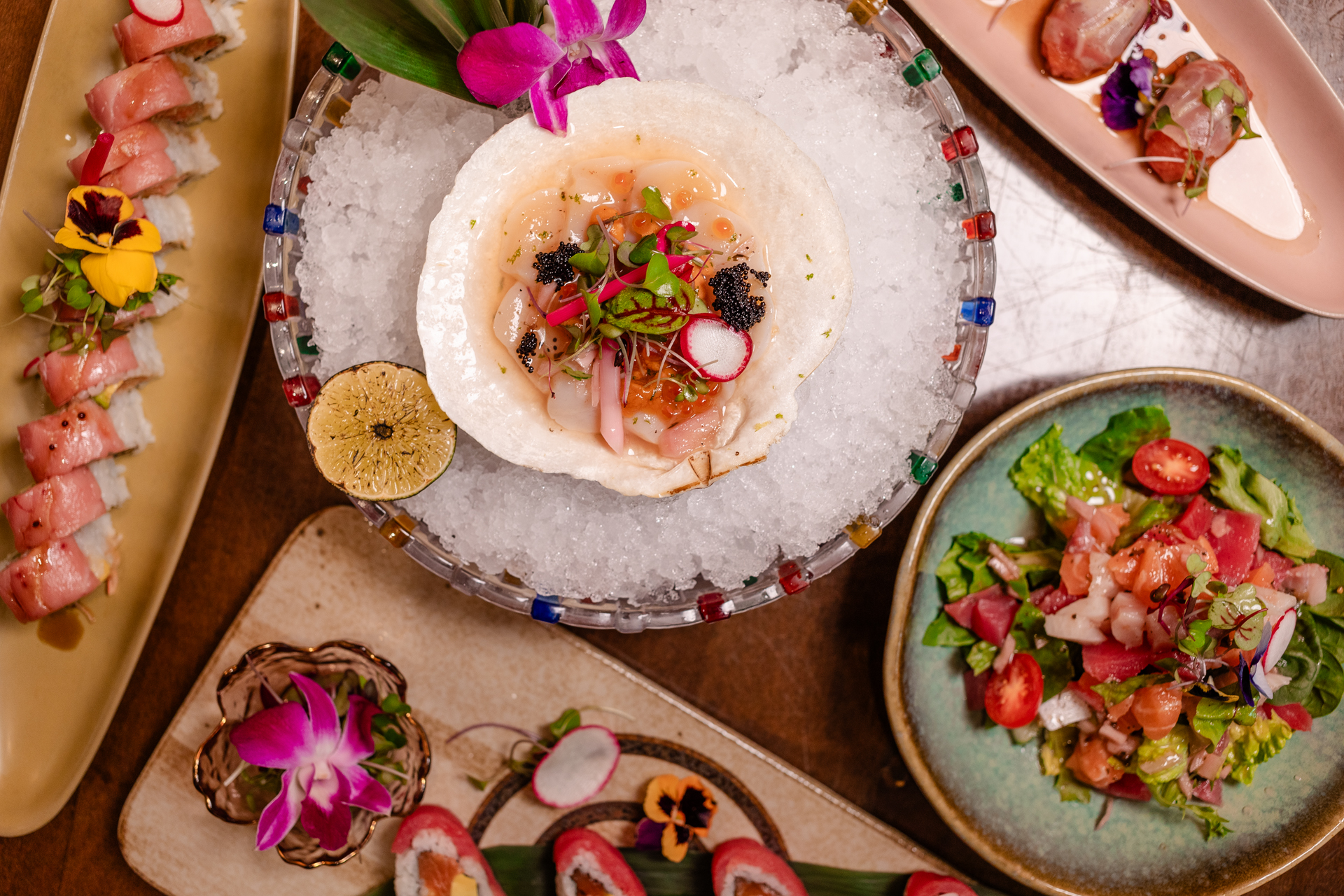 Overhead spread of sushi rolls, seafood crudo in a scallop shell over ice, and a tuna salad, garnished with radish and colorful edible flowers