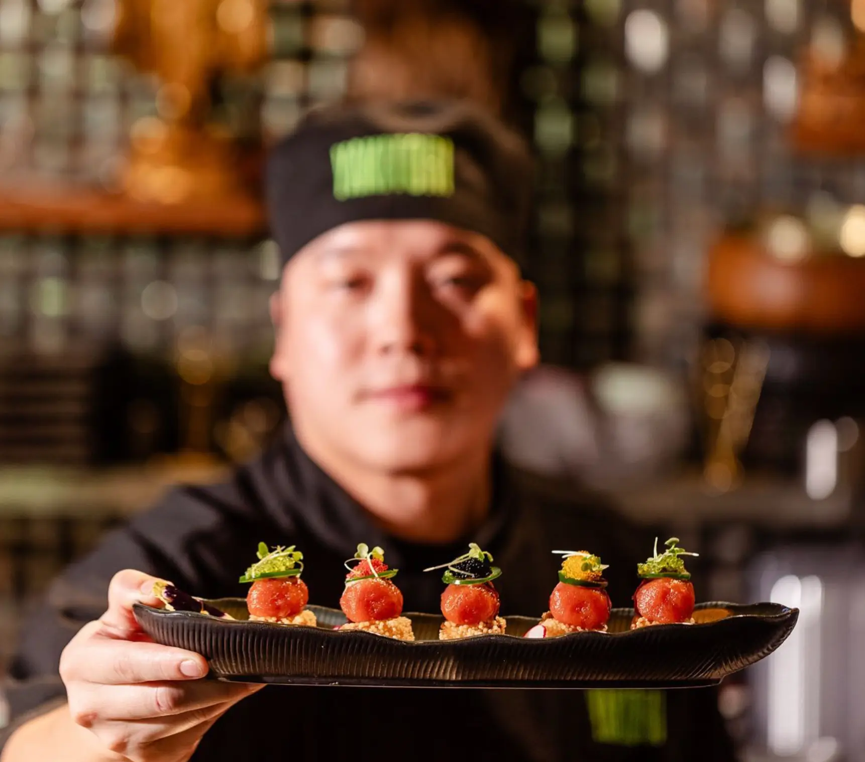 A chef in a black uniform holds forward a ridged black platter of five crispy rice bites topped with tuna tartare, jalapeño, microgreens and colorful roe