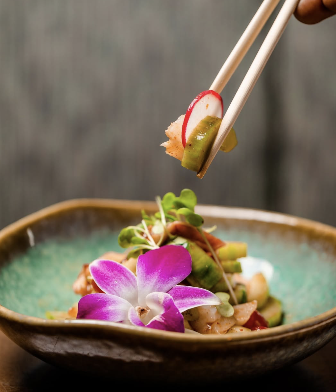 Chopsticks lifting a radish bite over a poke bowl garnished with a purple orchid