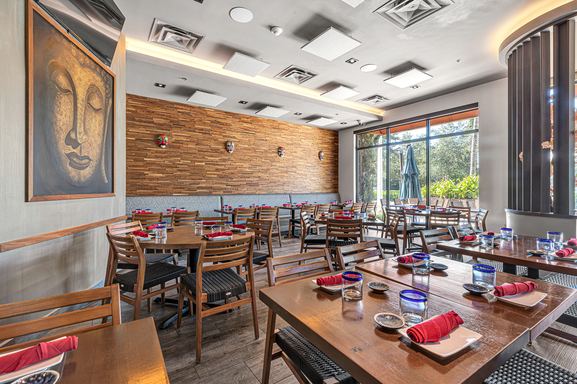 Sunlit dining room with polished wood tables set with rolled crimson napkins and blue-rimmed water glasses, a framed Buddha painting on the left wall and theatrical masks above a grey banquette