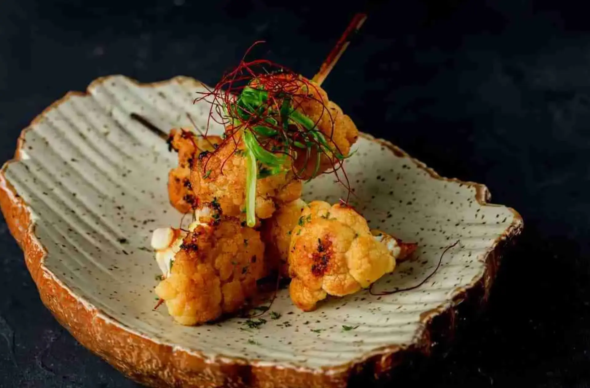 Grilled cauliflower florets on a seashell ceramic plate, garnished with scallion threads and a skewer